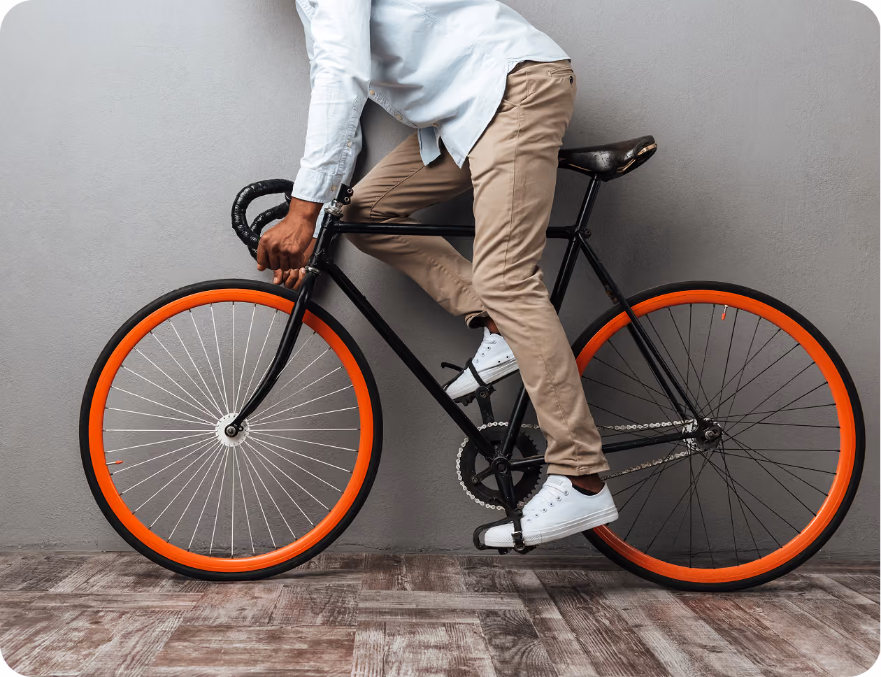 Person wearing khaki pants and white sneakers riding a black bicycle with orange rims indoors.