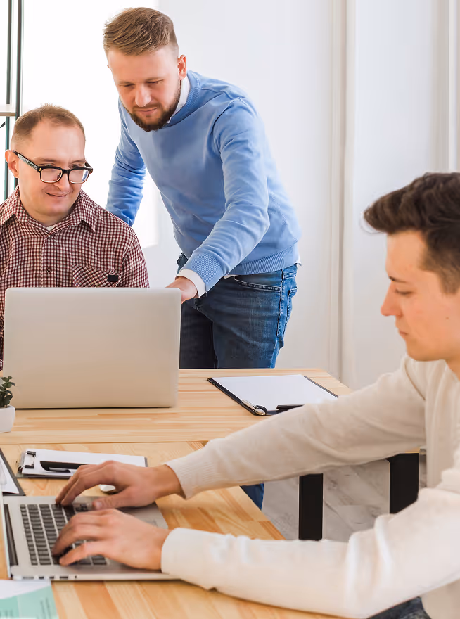Three men working together at a wooden table, two focused on a laptop and one typing on another laptop.