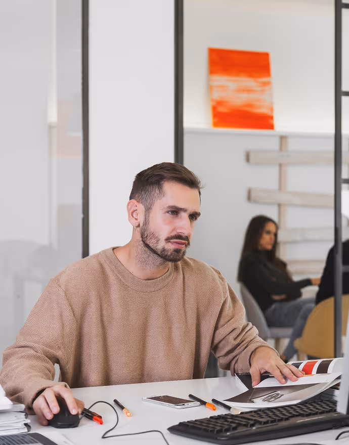 Man in a beige sweater working at a desk with a computer, holding a magazine and using a mouse in a modern office.