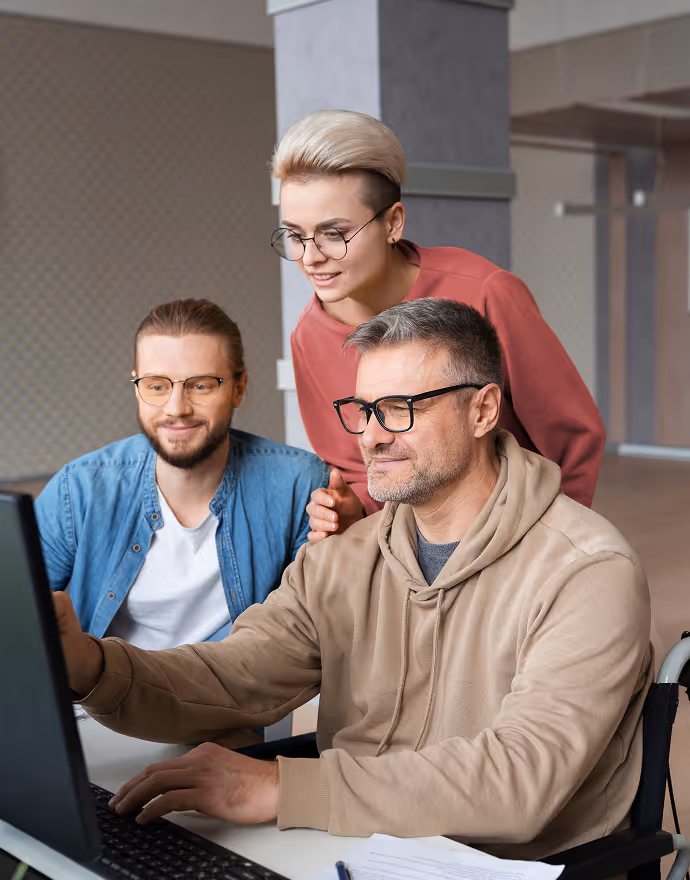 Three people wearing glasses working together on a computer in an office setting, one man in a wheelchair pointing at the screen.