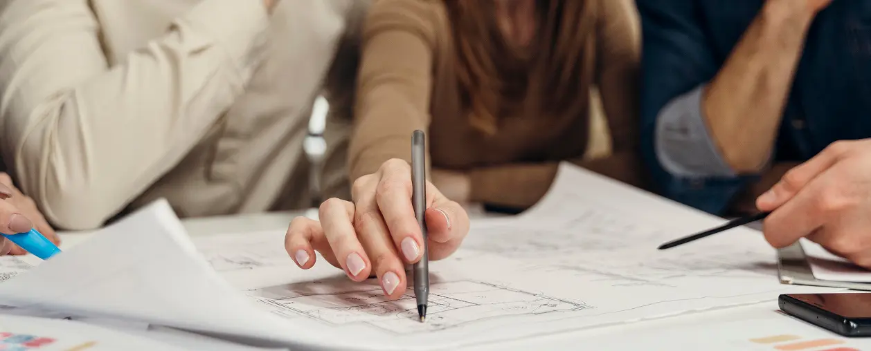 Close-up of a group of people collaborating over architectural blueprints, one hand holding a pen poised to draw.