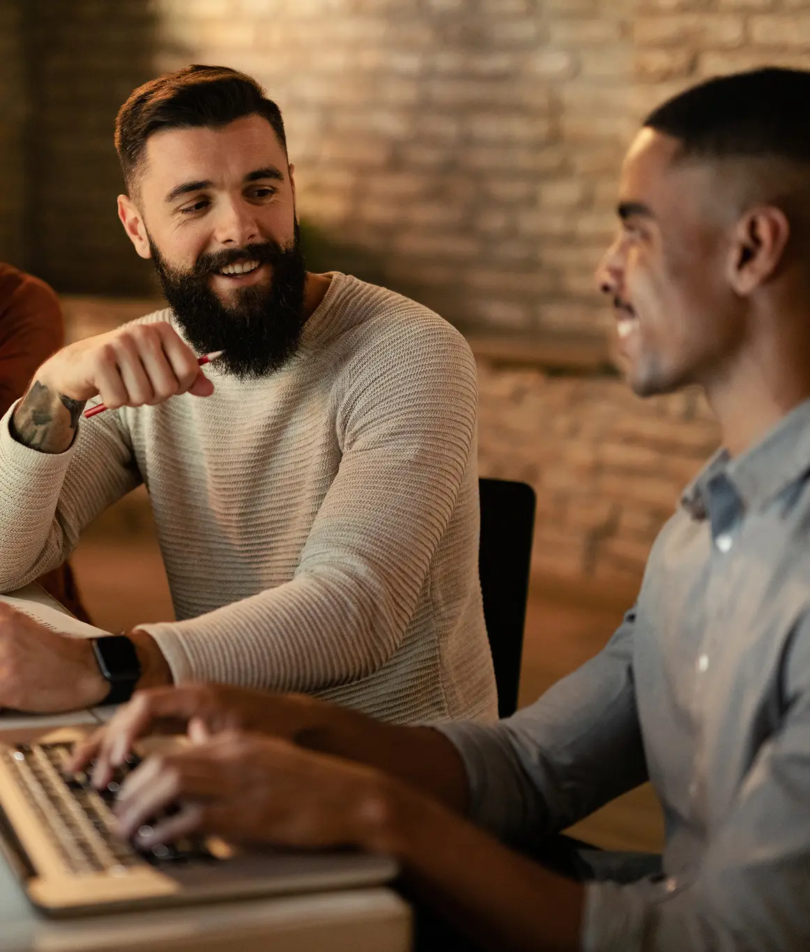 Two men smiling and working together on a laptop in a cozy, warmly lit room with a brick wall background.