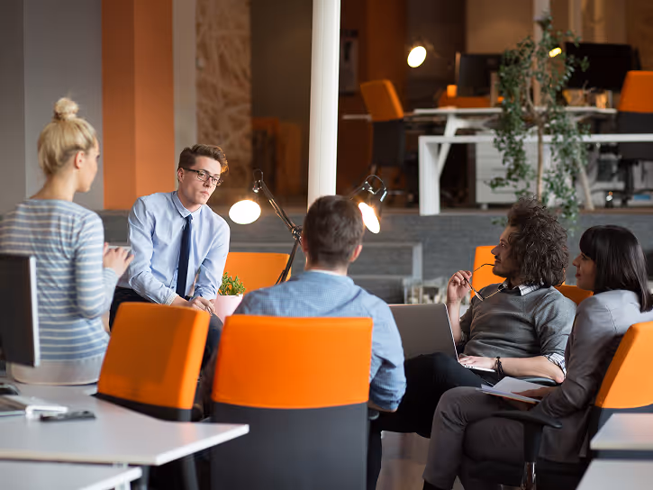 Group of five young professionals having a meeting in a modern office with orange chairs and desk lamps.
