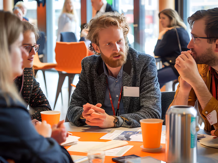 Group of four people engaged in a discussion around a table with papers and orange cups in a bright meeting room.