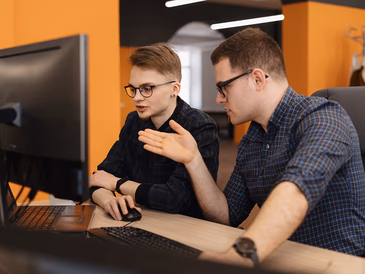 Two young men wearing glasses collaborating and discussing while looking at a computer monitor in an office with orange walls.