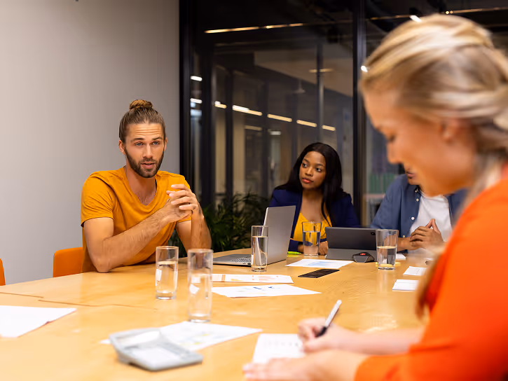 Four diverse colleagues at a conference table engaged in a business meeting with laptops, documents, and glasses of water.