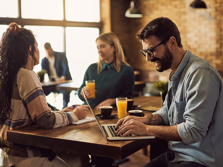 Three people sitting at a wooden table in a cozy cafe, one man working on a laptop smiling, two women talking with glasses of orange juice and coffee cups on the table.