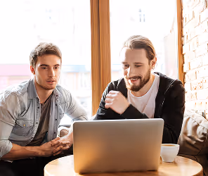 Two men sitting at a round table looking at a laptop in a bright room with large windows.