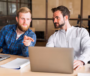 Two men sit at a table with laptops, one pointing at the screen while discussing with the other.