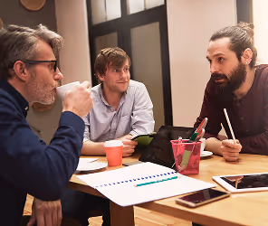 Three men having a discussion around a table with notebooks, tablets, and coffee cups.