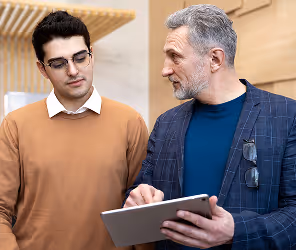 Two men discussing and looking at a digital tablet, one wearing glasses and a brown sweater, the other in a blue shirt and jacket.