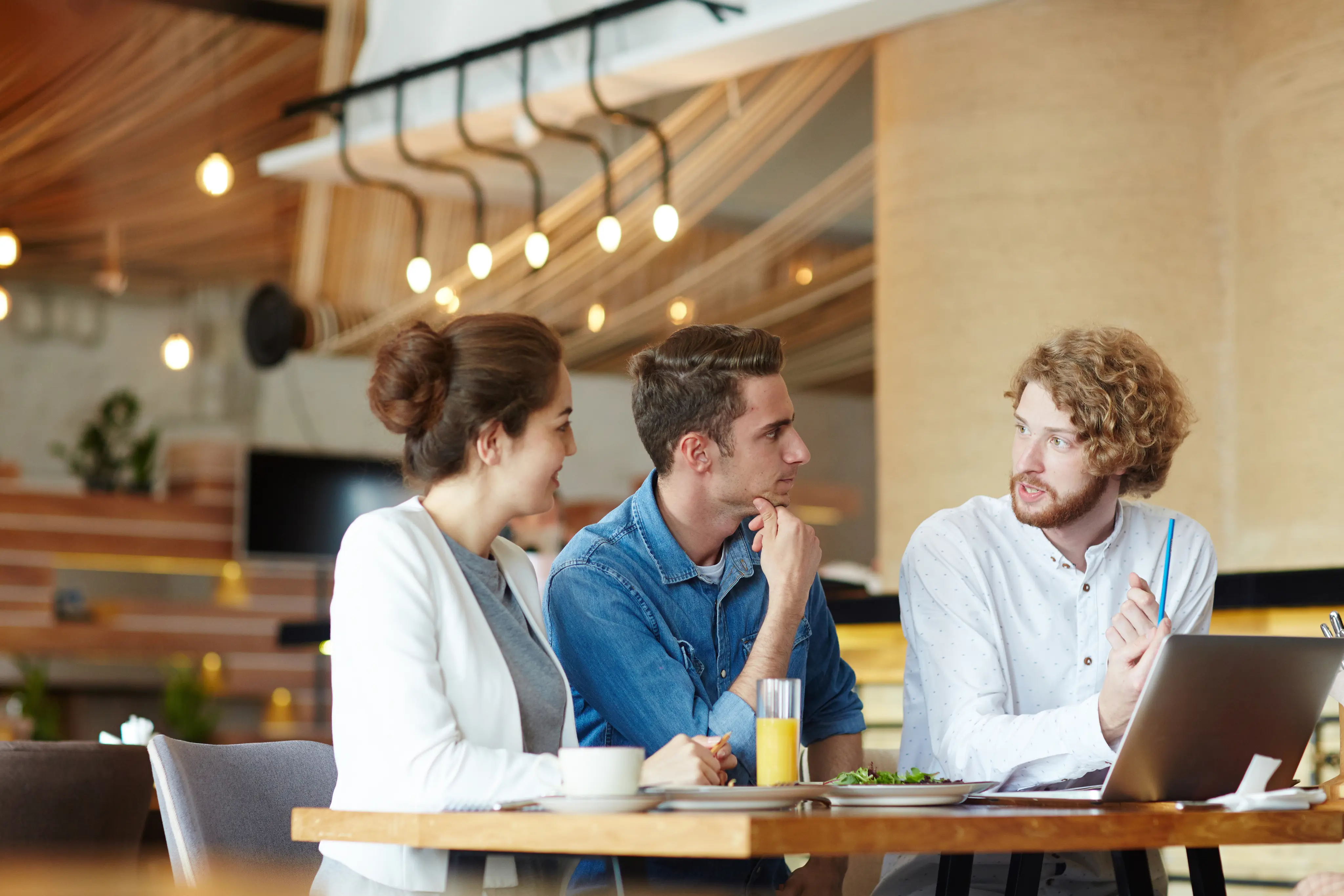 Three young professionals engaged in discussion at a cafe table with a laptop, drinks, and food.