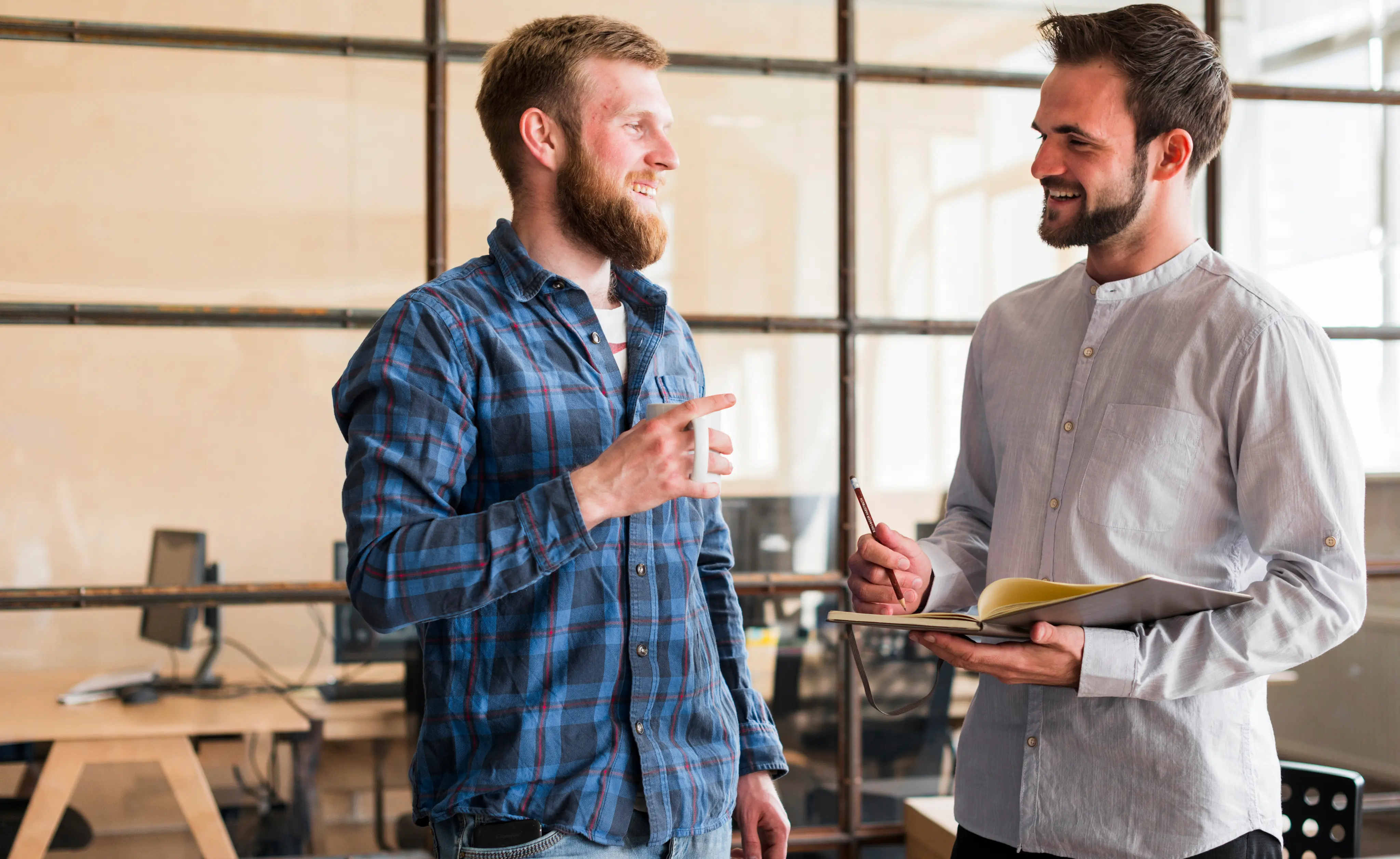 Two men having a conversation in an office, one holding a cup and the other holding a notebook and pencil.