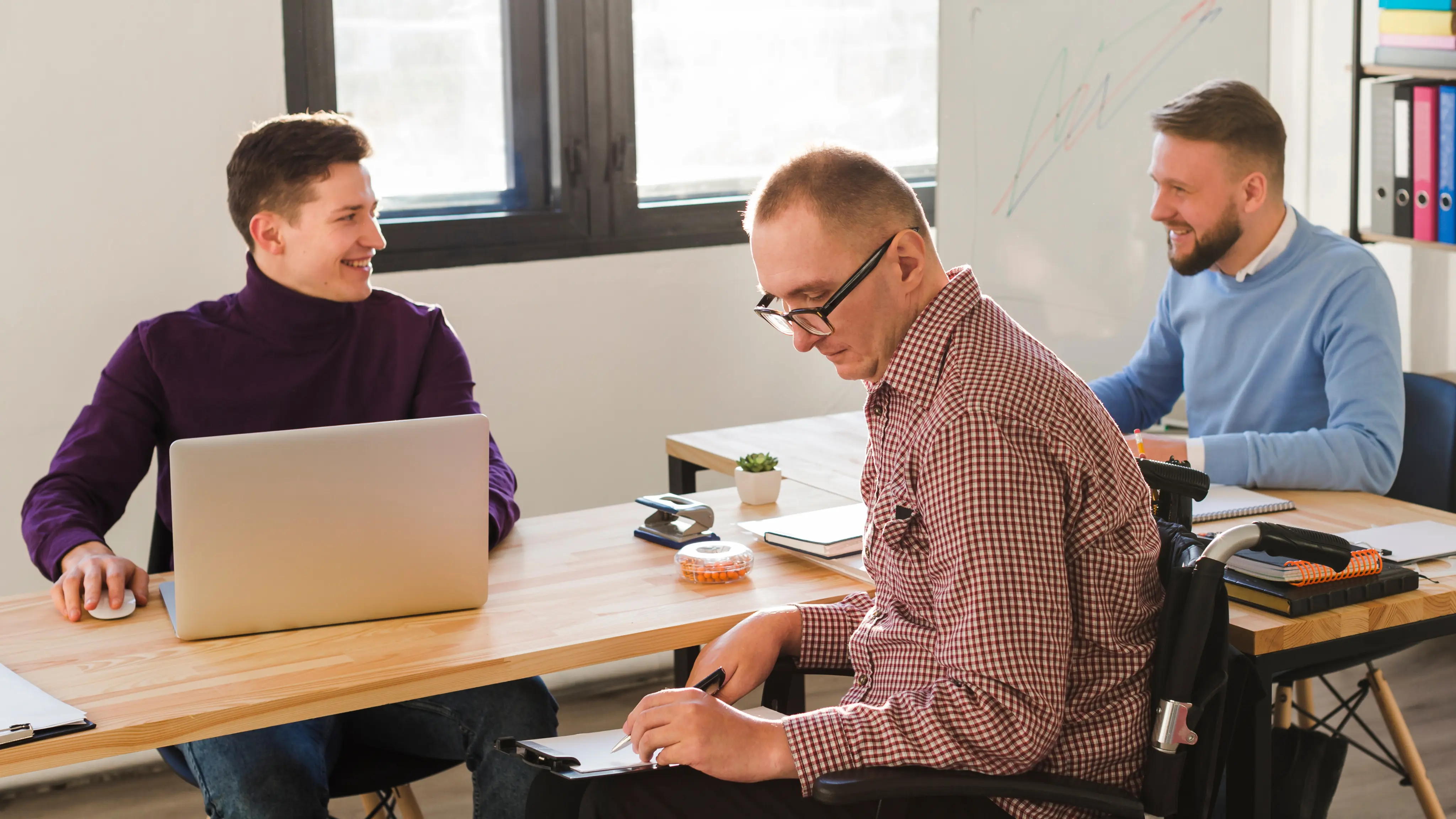 Three men in an office, one in a wheelchair writing on a clipboard and the other two smiling and using laptops.