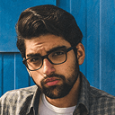 Man with dark hair and beard wearing glasses and a plaid shirt standing against a blue wooden background.